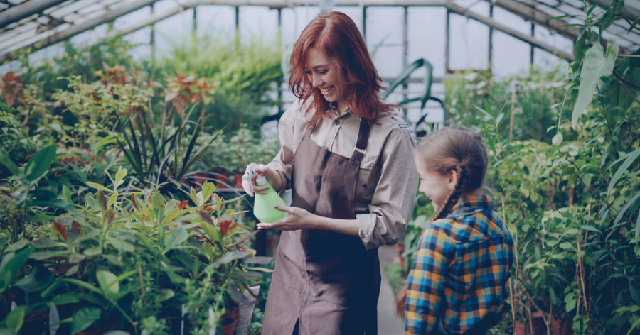 A woman with red hair is showing a young girl how to water plants with a spray bottle and other greenhouse gardening tips.