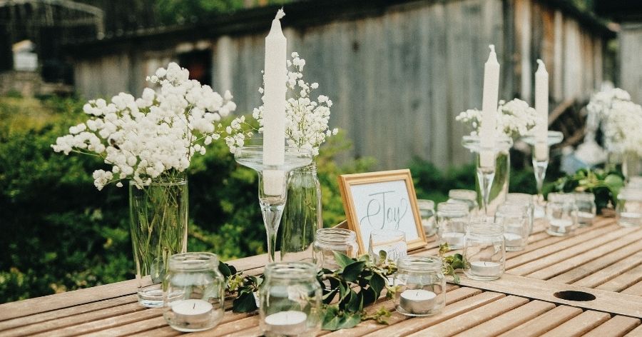 A DIY outdoor wedding table setting with taper and tea candles, white floral bouquets in glass vases, and green artificial foliage.