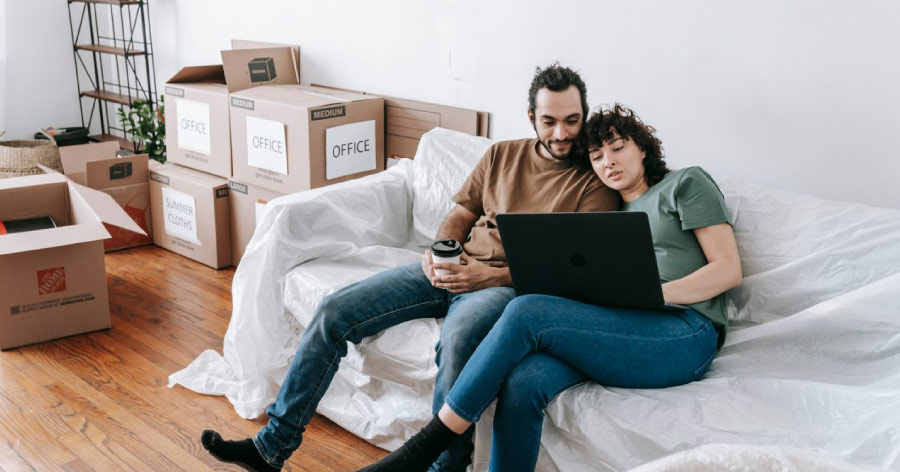 A couple is sitting on a covered couch beside stacks of moving boxes, using their laptop to book movers online and plan a moving timeline.
