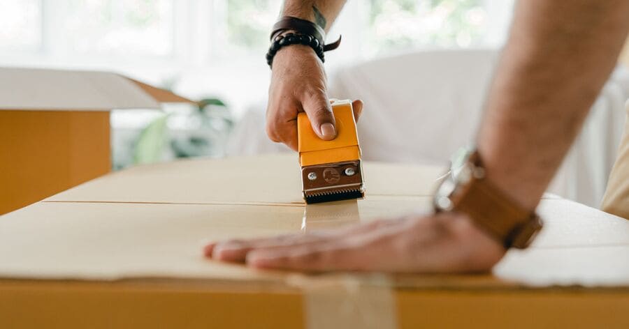 A man is sealing a cardboard moving box with packing tape while preparing to put things in storage during home renovations.