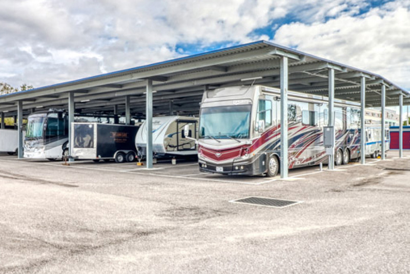 RVs are parked under a metal canopy in a storage parking lot.