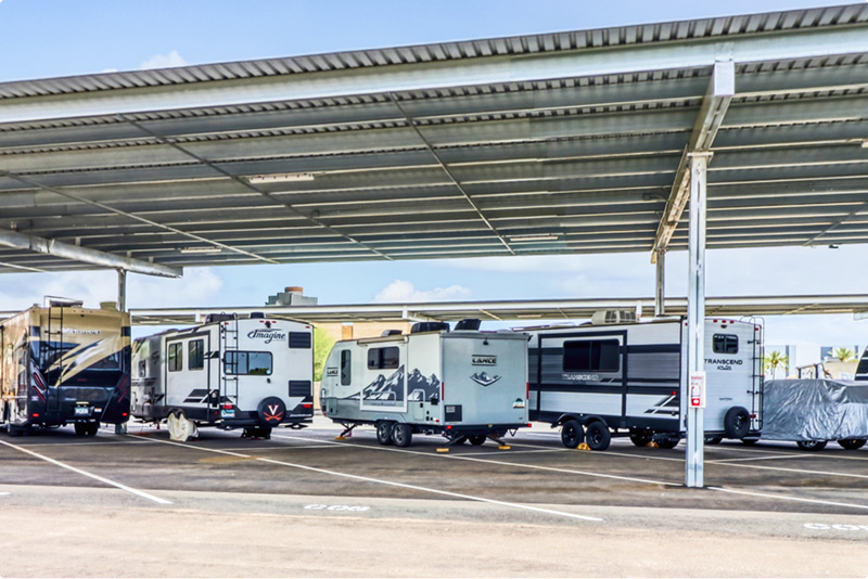 RVs are parked under a metal canopy in a storage parking lot.