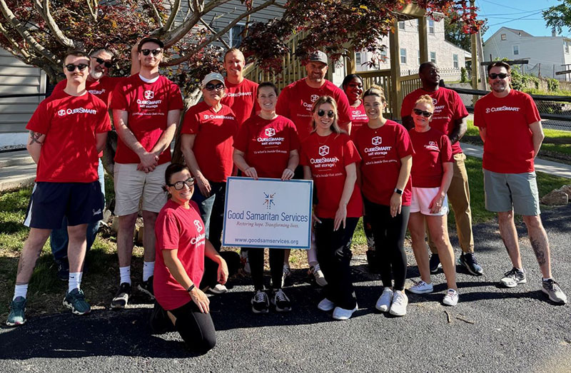 A large group of volunteers wearing matching red CubeSmart Self Storage t-shirts gathered outdoors under a tree, posing for a group photo around a sign that reads "Good Samaritan Services."
