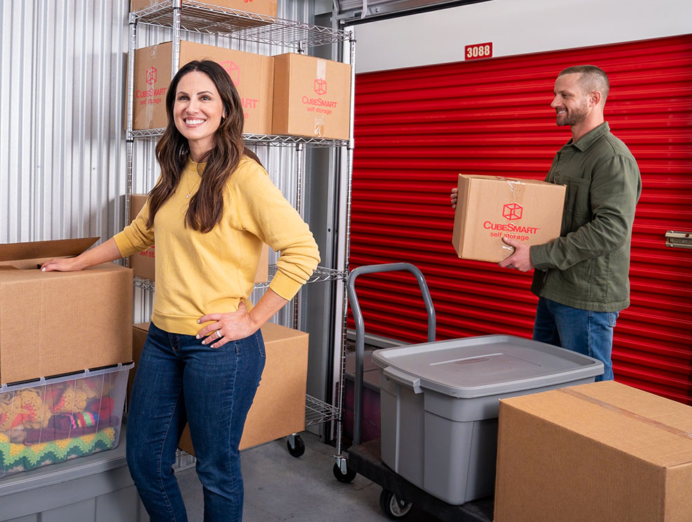 A woman in a yellow sweater and jeans poses inside an organized self-storage unit filled with cardboard boxes and plastic storage totes. In the background, a man carries a CubeSmart box toward the open red storage unit door.