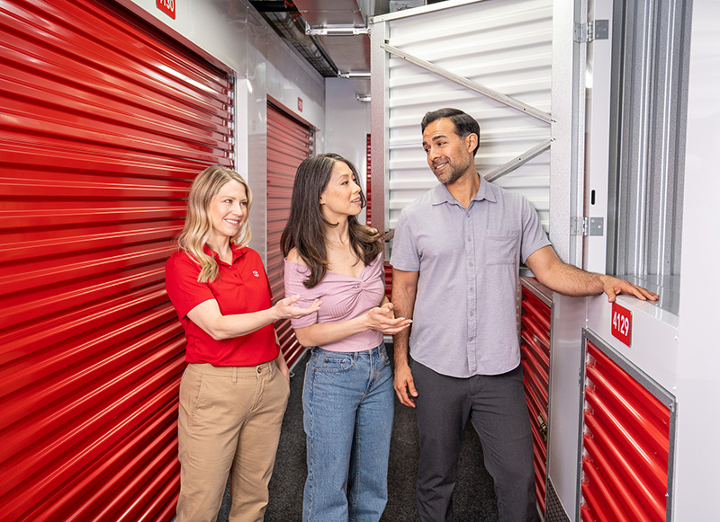 A self-storage facility employee in a red polo shirt showing an open storage locker to a couple in a brightly lit hallway.