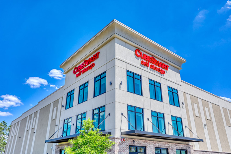 Low-angle shot of a multi-story CubeSmart Self Storage building with beige siding, large windows, black awnings, and red signage under a clear blue sky.