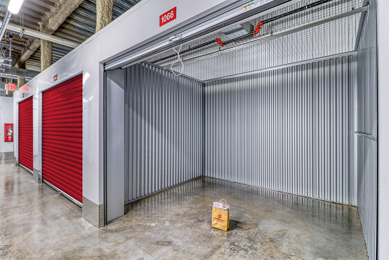 Brightly lit hallway in an indoor storage facility featuring an open unit with corrugated metal walls and a small paper bag on the clean concrete floor, alongside closed red unit doors.