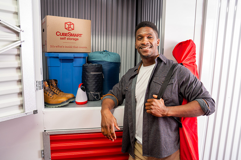 A man smiling at the camera while leaning against an open upper storage locker containing hiking boots, sleeping bags, a lantern, a blue plastic tote, and a CubeSmart box.