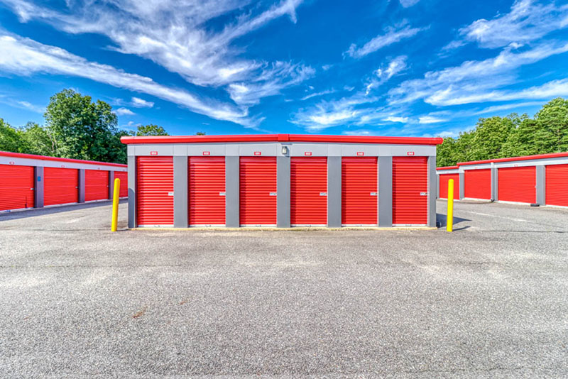Row of outdoor, drive-up self-storage units with bright red roll-up doors, gray trim, and yellow safety bollards set on a paved asphalt lot.